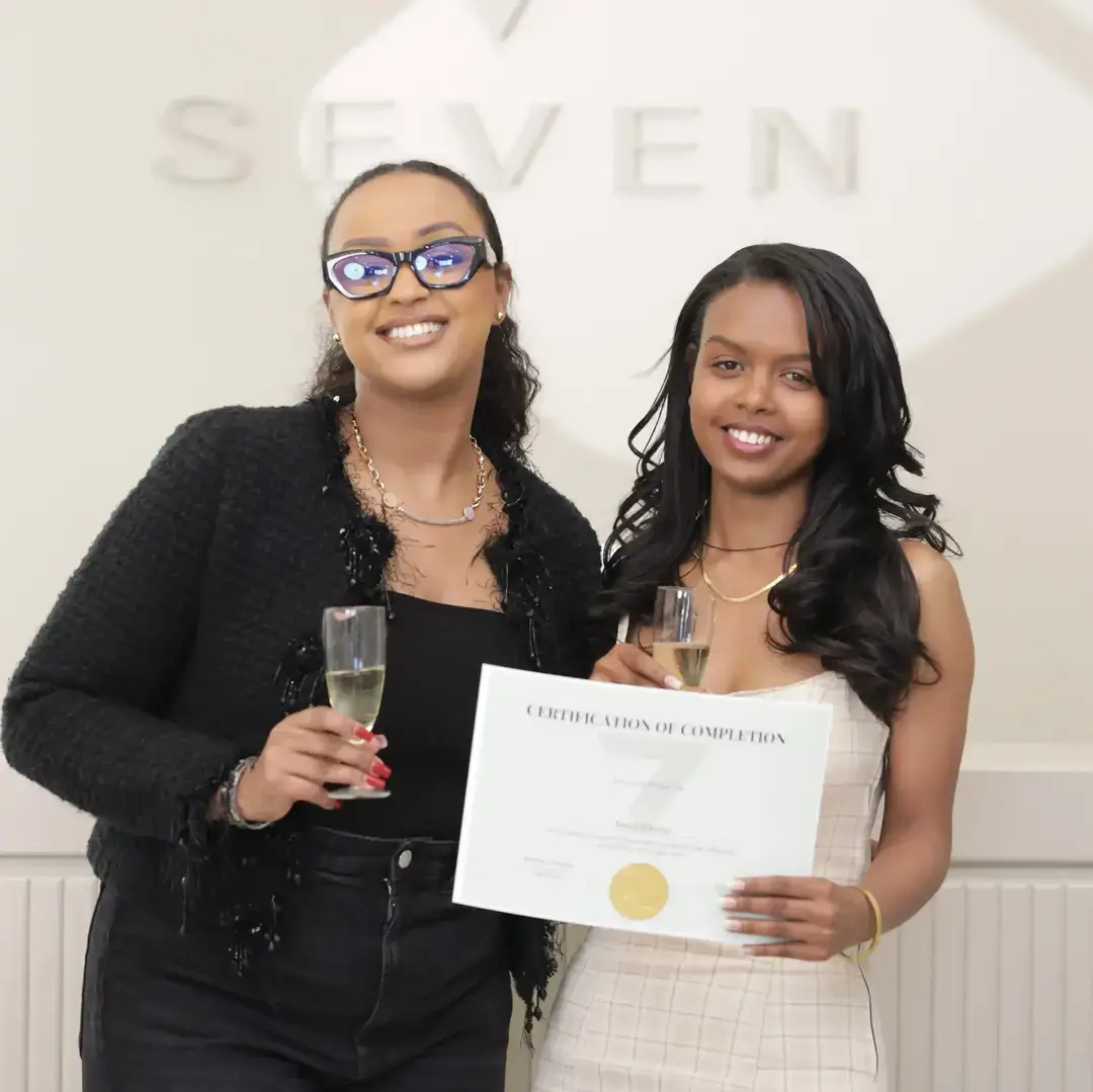 Permanent Makeup Certification at Seven Aesthetics: Teacher Betlehem Bereket with student Ruth Alemayehu holding her certificate of completion, celebrating with champagne after completing the course.
